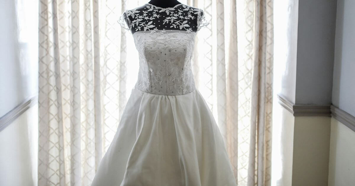 A beautifully preserved white wedding dress displayed on a mannequin in front of softly lit curtains. The dress features intricate lace and floral details on the bodice, highlighting elegance and care in preservation.