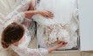 A woman arranges a white wedding dress with intricate details inside a box while wearing a white lace robe. The scene, viewed from above, features STEP 4: Additional Items.